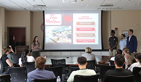 Students give a presentation in front of a large screen with a slide that says Tim Hortons and includes other information