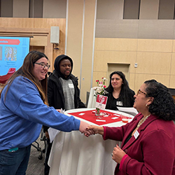 A student shakes hands with a staff member. 2 other students stand alongside them.