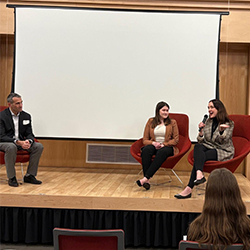 A woman sits in a chair on stage and speaks. 2 other professionals sit alongside her.