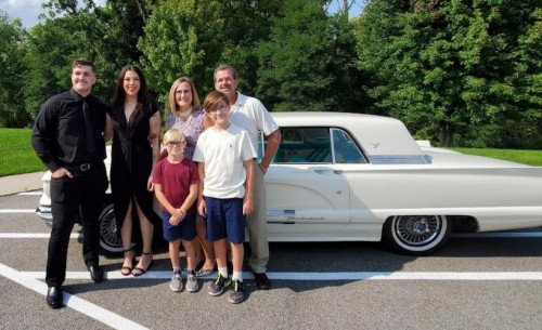 4 adults and 2 kids stand and smile in front of a car