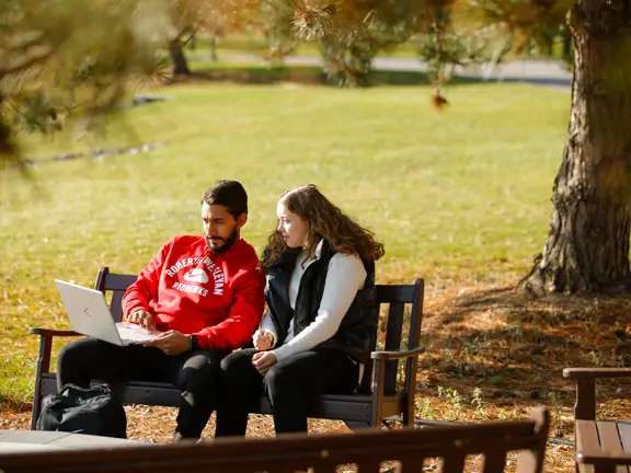 2 students sit on a bench and look at a laptop together