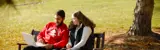 2 students sit on a bench and look at a laptop together