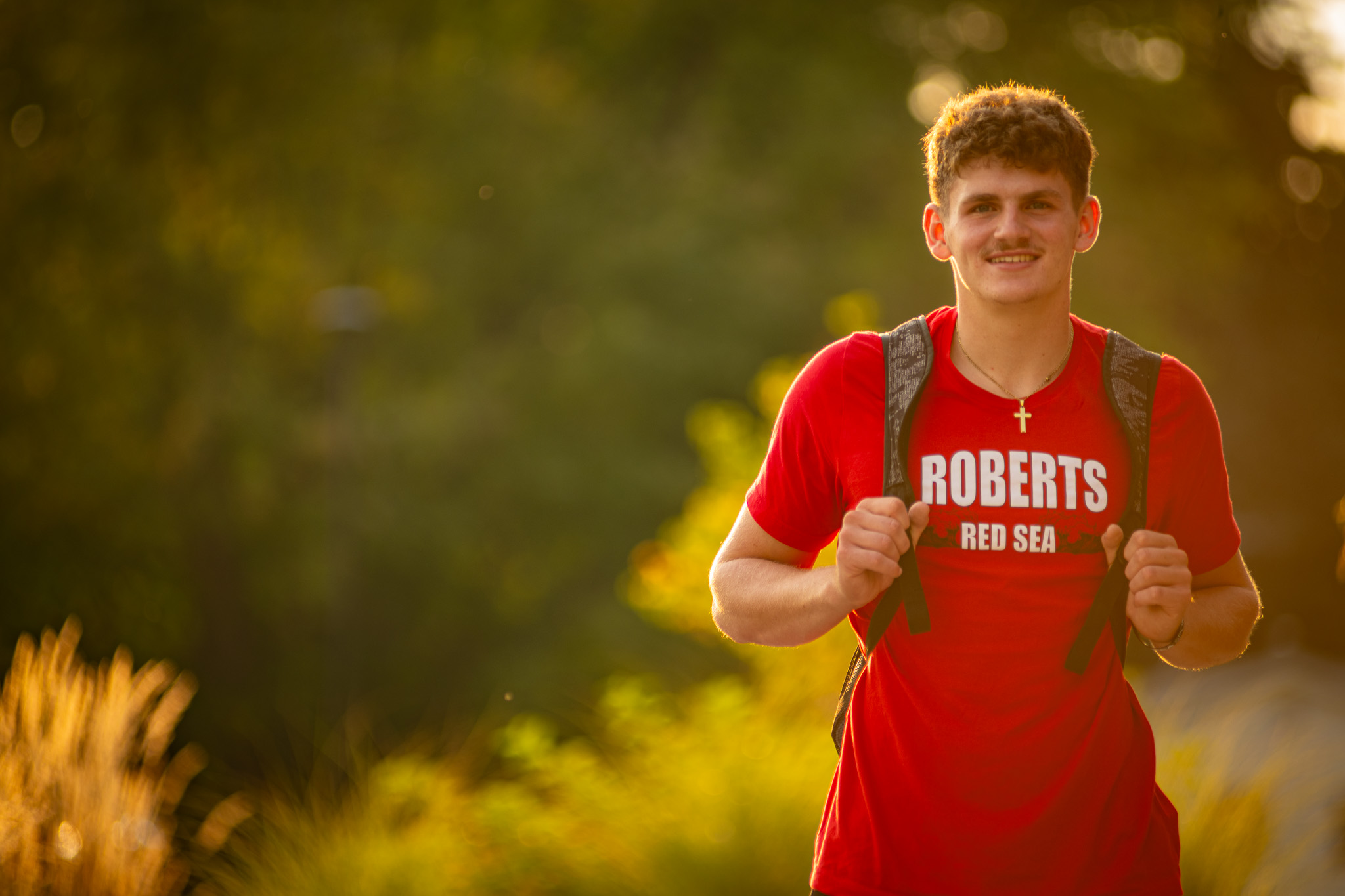 A young man stands on campus while wearing a Roberts shirt and smiling