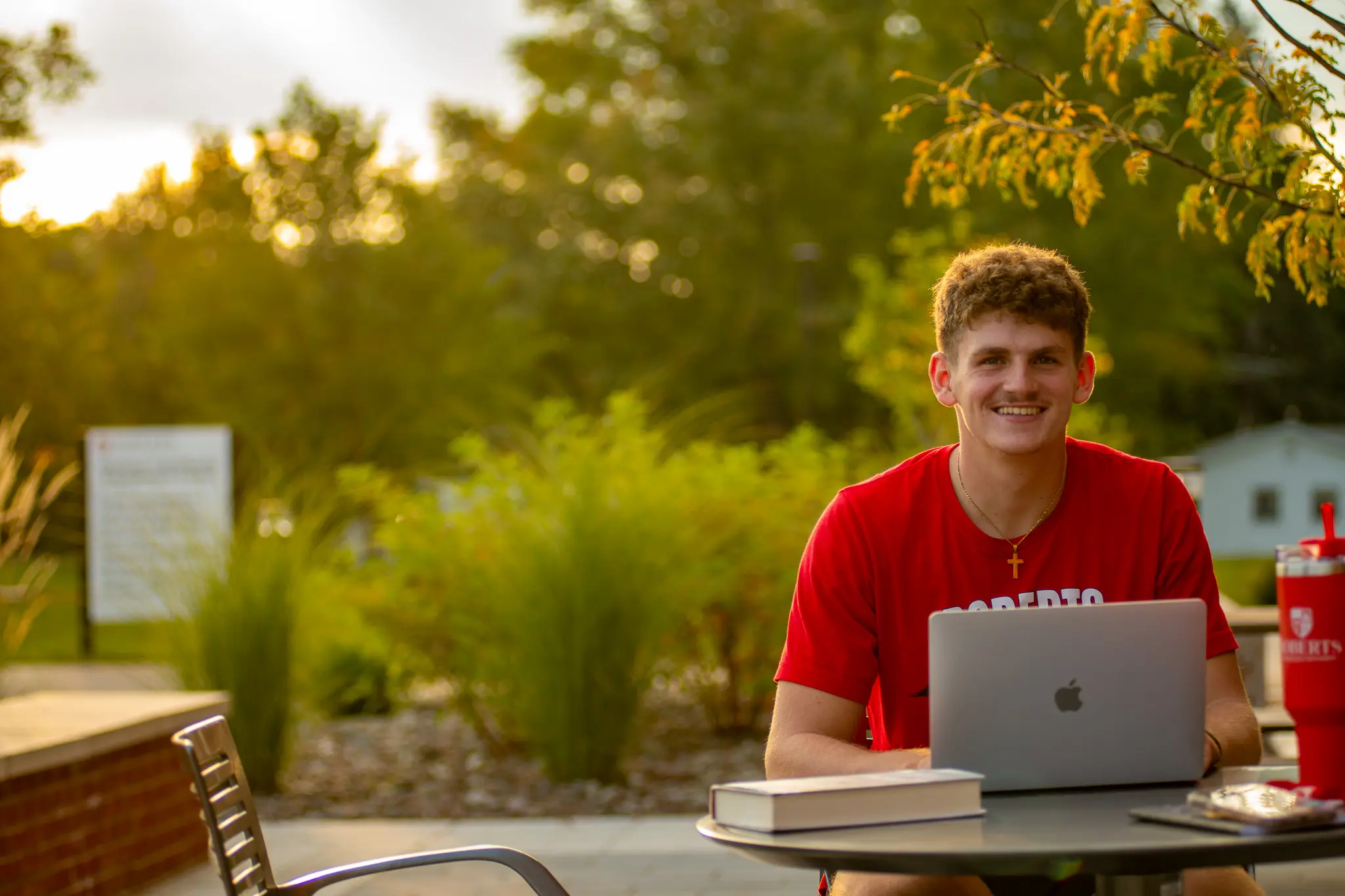 A young man sits with a laptop open and smiles